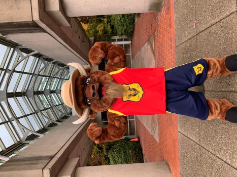 A bison mascot dressed in a basketball uniform, wearing a red RCMP basketball jersey and blue basketball shorts and a brown Stetson hat standing in front of the Surrey RCMP building.